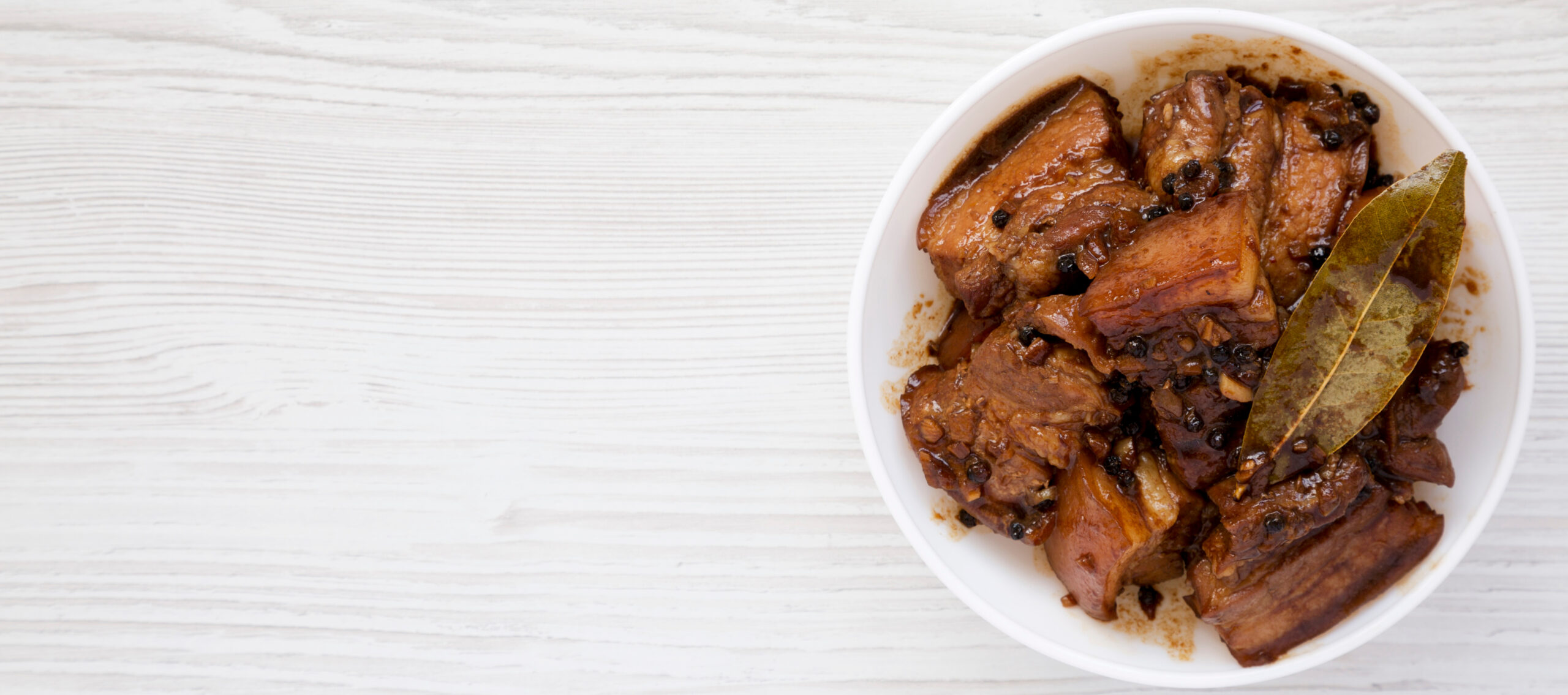 Homemade Filipino Adobo Pork in a white bowl over white wooden background, top view. Flat lay, from above, overhead. Space for text.